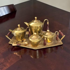 May include: A vintage brass tea set including a teapot, creamer, sugar bowl, and serving tray. The set features wooden handles and a classic design. The tray is approximately 30 cm long.