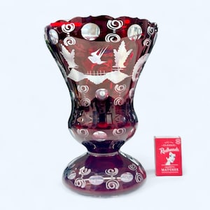 May include: A deep red glass vase with a flared top and a pedestal base. The vase has etched white patterns of birds, trees, and swirls. A small box of Redheads matches is next to the vase.