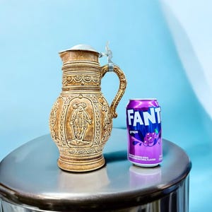 May include: A brown ceramic beer stein with a pewter lid and a detailed design of a man in a traditional costume. The stein is sitting on a silver table next to a purple can of grape soda labelled "Fanta".