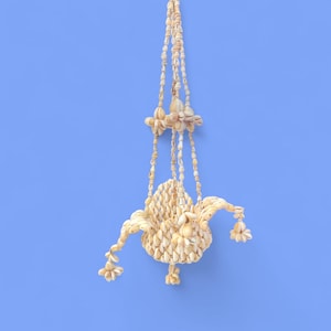May include: A hanging ornament made from seashells, suspended by beaded strands. The central form is a rounded shape with extended, curved elements, and adorned with clusters of shells. The background is a solid blue.