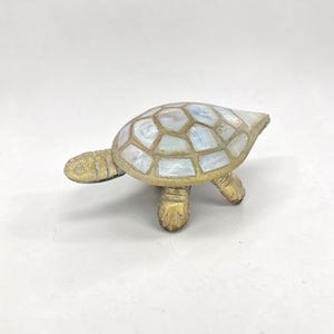 May include: A decorative turtle figurine featuring a textured, antique gold-toned metal head, legs, and tail. The shell is crafted from inlaid, iridescent white mother-of-pearl segments arranged in a geometric pattern, creating a shimmering effect.