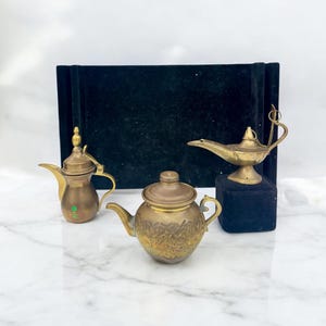 May include: Three antique brass decorative items: a teapot, a coffee pot, and a genie lamp. The teapot has an embossed floral design. The coffee pot has a green emblem. The genie lamp sits on a black base. All items are on a white marble surface.