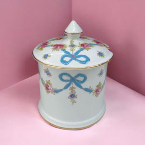May include: A white ceramic jar with a lid, decorated with pink roses, blue ribbons, and gold trim. The jar features a conical finial and a matching floral design on the lid. A decorative item for the home.