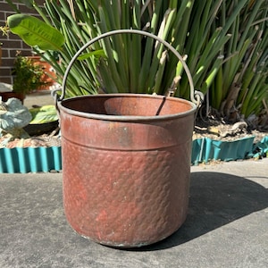 May include: A hammered copper planter with a wire handle. The planter is a reddish brown colour and has a rustic look.