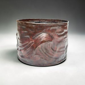 May include: A handmade ceramic pot with a cylindrical shape and a textured, wave-like design. The exterior is a deep reddish-brown colour with tonal variations. The open top and base are visible. Likely a decorative item or small container.