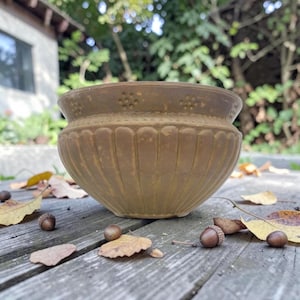 May include: A bronze-coloured, antique-style planter with a wide, rounded shape and vertical ribbed detailing. The rim features a decorative dotted pattern. Acorns and fallen leaves rest on the wooden surface around the planter.
