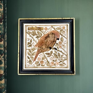 May include: A square framed wall art piece featuring a brown bird perched on a branch amidst floral foliage. The artwork is set against a white background with gold accents and the text BULLFINCH printed in a rectangular box at the top left.