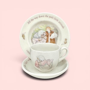 May include: A ceramic tea cup and saucer set with a matching bowl featuring illustrations of a hedgehog in a bonnet and a person in a brown coat. The text reads All the way down the path little animals came Tiggy-winkle and a mouse.