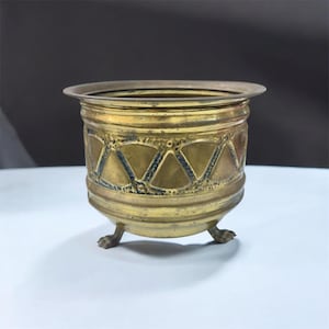 May include: A vintage brass planter with a geometric pattern and three claw feet. The planter has a wide rim and a cylindrical body. The brass has a warm, golden hue, and the geometric design is accented with dark blue.