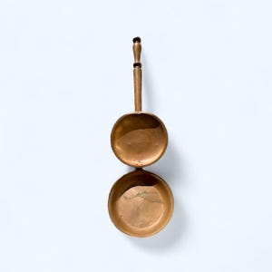 May include: A vintage copper ladle with a long handle and a round bowl. The ladle is made of copper and has a warm, metallic colour. The handle has a decorative design.