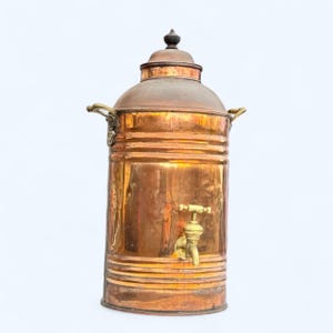 May include: A vintage copper urn with a brass tap and handles. The urn features a domed lid with a decorative finial. The copper has a warm, reddish-brown hue, and the brass fittings add a contrasting golden colour.