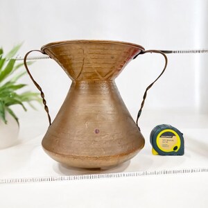 May include: A copper-coloured vase with two twisted handles. The vase has a wide top and a rounded base. A black and yellow tape measure is next to the vase. A green plant in a white pot is in the background.