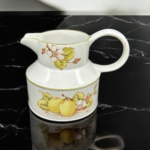 May include: White ceramic creamer with a floral design featuring yellow and green leaves, pink flowers, and yellow fruit. The creamer has a handle and a rounded body.