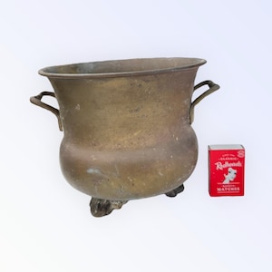 May include: A brass planter with two handles and three feet. The planter is oval shaped and has a worn, aged look. A box of matches is in the background for scale.
