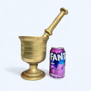 May include: A vintage brass mortar and pestle set with a cylindrical bowl and a long, rounded pestle. A purple and blue Fanta grape soda can is next to the mortar. The mortar and pestle are on a white background.