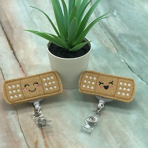 May include: Two tan felt bandage badge reels with smiling faces and white dot details. The badge reels have a clear plastic retractable cord and clip. A small potted plant is in the background.