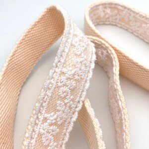 May include: Two beige cotton ribbon trims with white embroidered floral designs. The ribbon trims are woven and have a soft, textured feel.