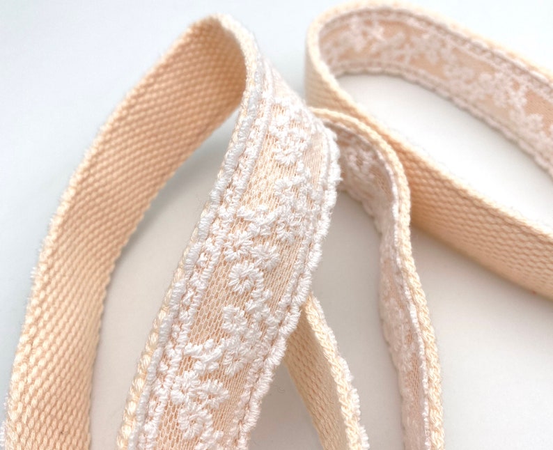 May include: Two beige cotton ribbon trims with white embroidered floral designs. The ribbon trims are woven and have a soft, textured feel.