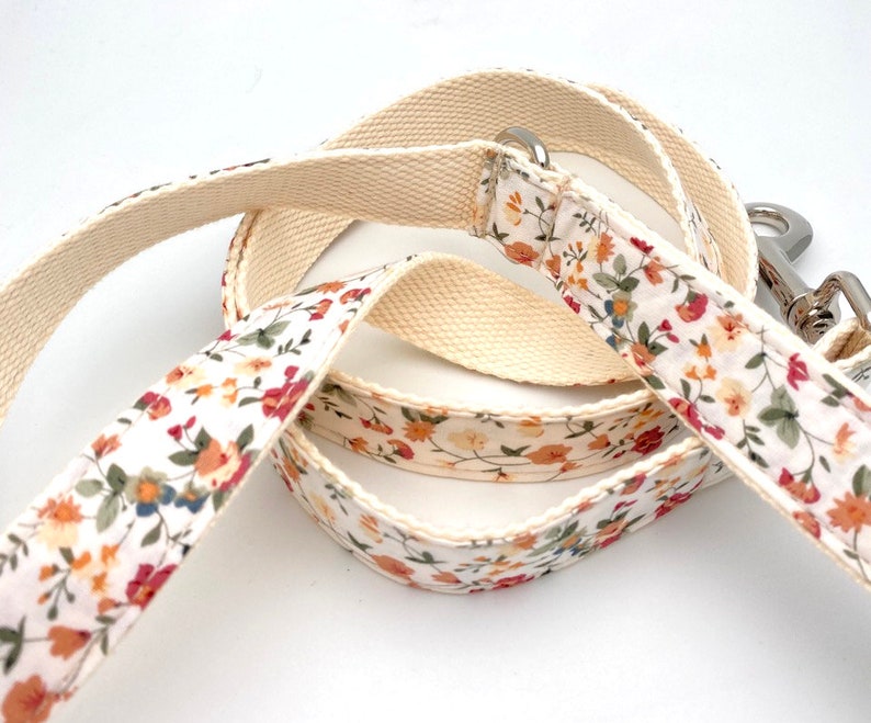 May include: A white cotton dog leash with a floral pattern in shades of orange, red, and green. The leash has a silver metal clip.