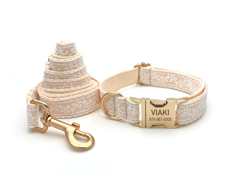 May include: A beige dog collar and lead set with a gold buckle and a personalised name tag that reads "VIAKI 876-987-0000". The collar and lead feature a delicate white floral pattern.