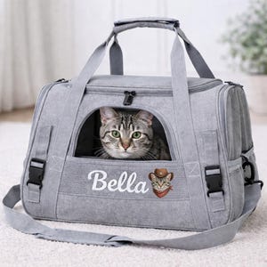 May include: A gray cat carrier with a cat peeking out of the front window. The carrier has a shoulder strap and handles. The name "Bella" is embroidered on the front, along with a cat wearing a cowboy hat and bandana.