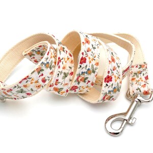 May include: A white cotton dog leash with a floral print featuring red, orange, and yellow flowers. The leash has a silver metal clip.