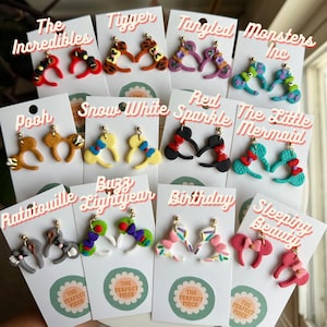 May include: A collection of handmade earrings featuring various character designs. Each pair is attached to a white card with the text "The Perfect Piece". Designs include "The Incredibles", "Tigger", "Tangled", "Monsters Inc", "Pooh", "Snow White", "Red Sparkle", "The Little Mermaid", "Ratatouille", "Buzz Lightyear", "Birthday", and "Sleeping Beauty".