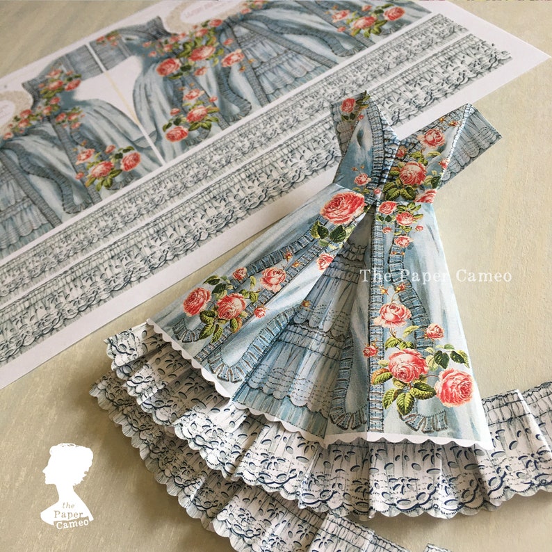 May include: A paper craft project featuring a light blue dress with a floral rose design. The dress has a layered skirt with white lace trim. Additional printed dress patterns and lace trim are visible.