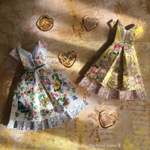 May include: Two origami dresses, one white with butterfly and floral patterns, the other yellow with roses. Gold heart-shaped wax seals and a bee seal are scattered around the dresses. The Paper Cameo logo is in the bottom right corner.