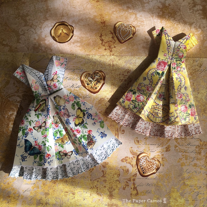 May include: Two origami dresses, one white with butterfly and floral patterns, the other yellow with roses. Gold heart-shaped wax seals and a bee seal are scattered around the dresses. The Paper Cameo logo is in the bottom right corner.