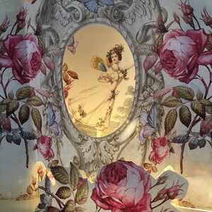 May include: A vintage papercraft with a delicate floral design featuring pink roses, green leaves, and butterflies. A framed image of a woman in a white dress with butterfly wings is visible through a cutout in the papercraft.
