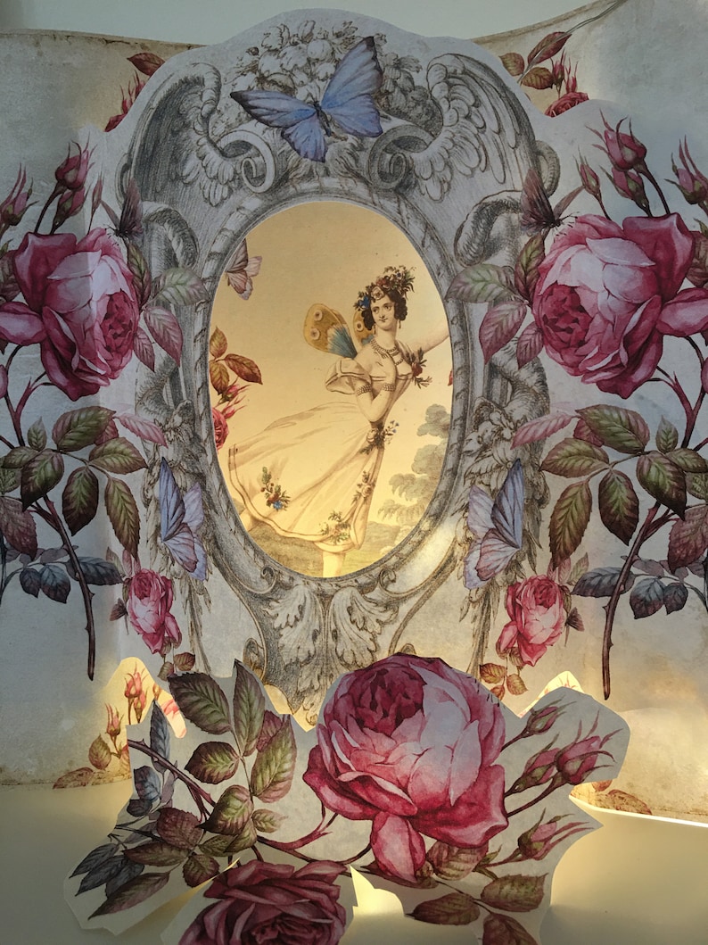 May include: A vintage papercraft with a delicate floral design featuring pink roses, green leaves, and butterflies. A framed image of a woman in a white dress with butterfly wings is visible through a cutout in the papercraft.
