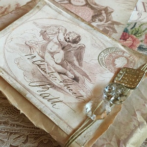 May include: A vintage-style gift box wrapped in brown paper with a delicate lace overlay. The box features a drawing of a cherub with wings and the text "It's  a  Love  Gift  From  the  Heart".