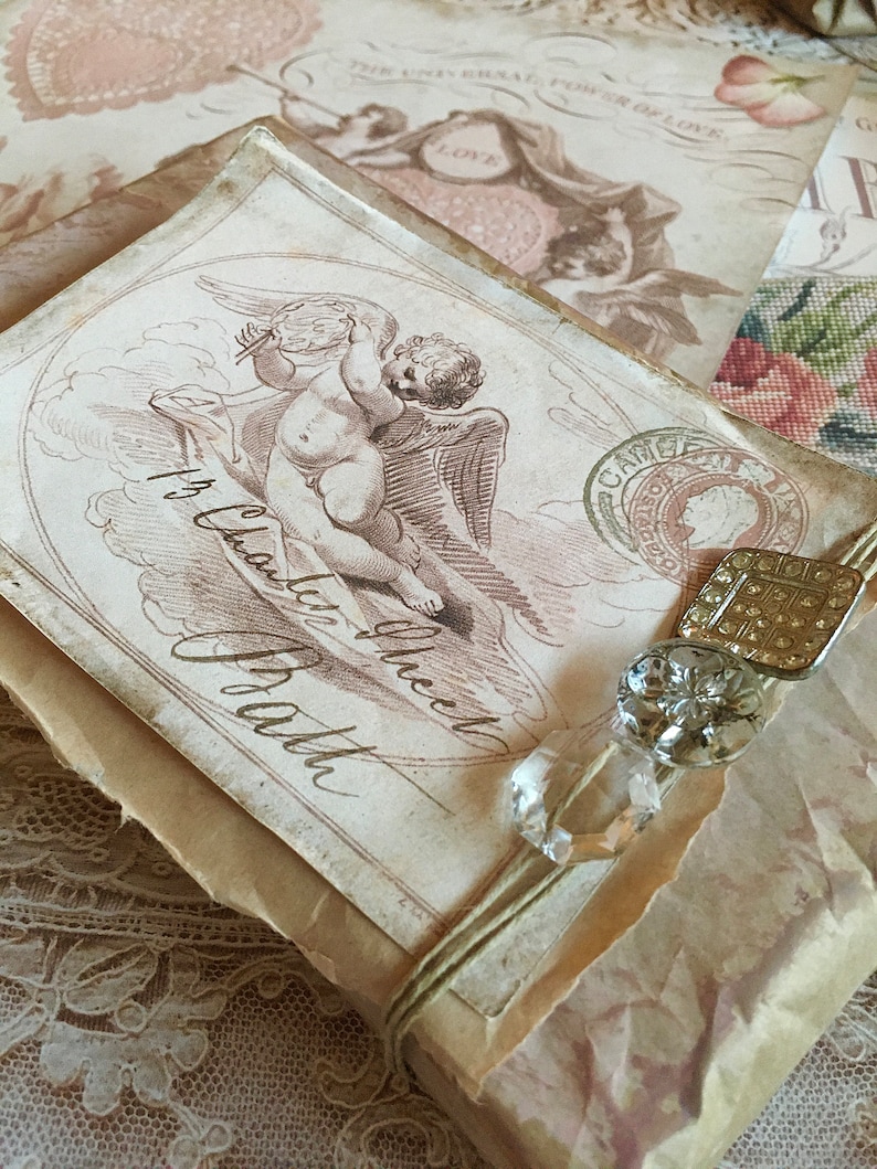 May include: A vintage-style gift box wrapped in brown paper with a delicate lace overlay. The box features a drawing of a cherub with wings and the text "It's  a  Love  Gift  From  the  Heart".