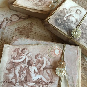 May include: A stack of vintage-style boxes with a distressed, aged look. Each box features a different image of cherubs and angels, with some boxes adorned with decorative buttons and twine. The boxes are decorated with a faded, antique aesthetic.