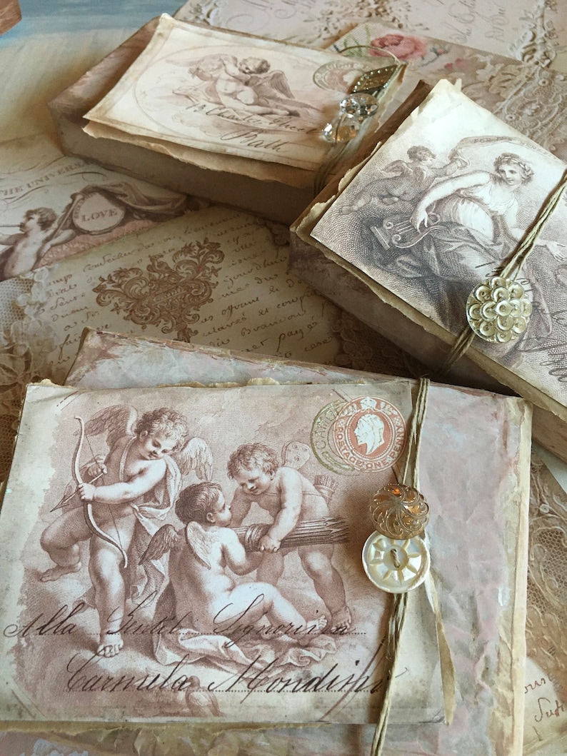 May include: A stack of vintage-style boxes with a distressed, aged look. Each box features a different image of cherubs and angels, with some boxes adorned with decorative buttons and twine. The boxes are decorated with a faded, antique aesthetic.