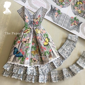 May include: A paper dress with a gray and white floral and butterfly pattern. The dress has a V-neck, short sleeves, and a layered skirt with a scalloped lace trim. The Paper Cameo logo is visible.