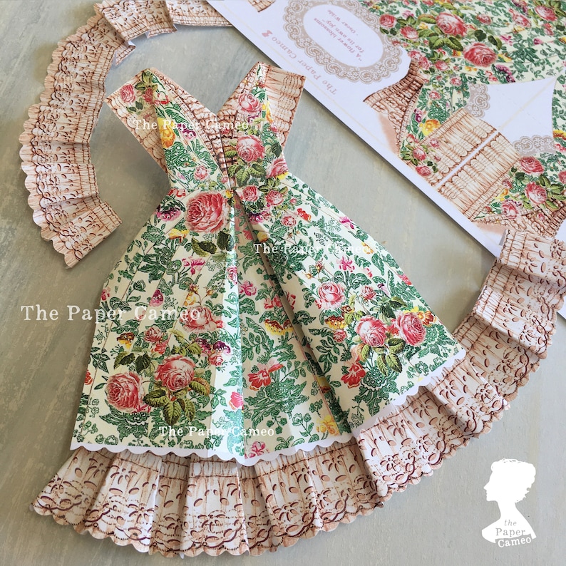 May include: A paper dress with a floral pattern of red roses and green leaves. The dress has a scalloped hem and is accompanied by matching paper lace trim. The text "The Paper Cameo" is visible on the dress.
