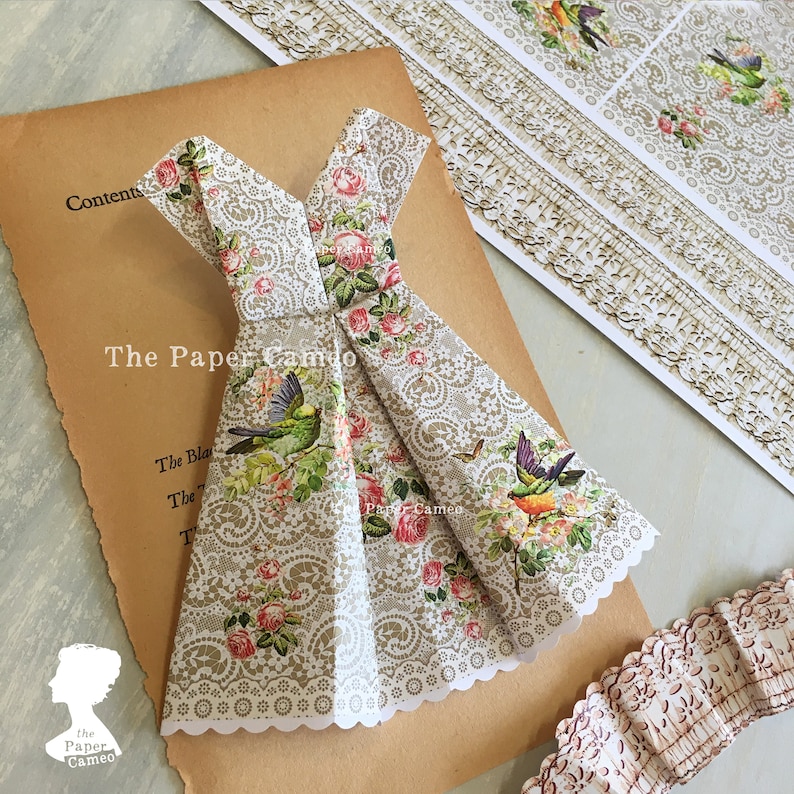 May include: A paper dress with a floral and bird pattern, featuring pink roses and green hummingbirds, is displayed on a vintage book page. The dress has a lace-like design and the text "The Paper Cameo".
