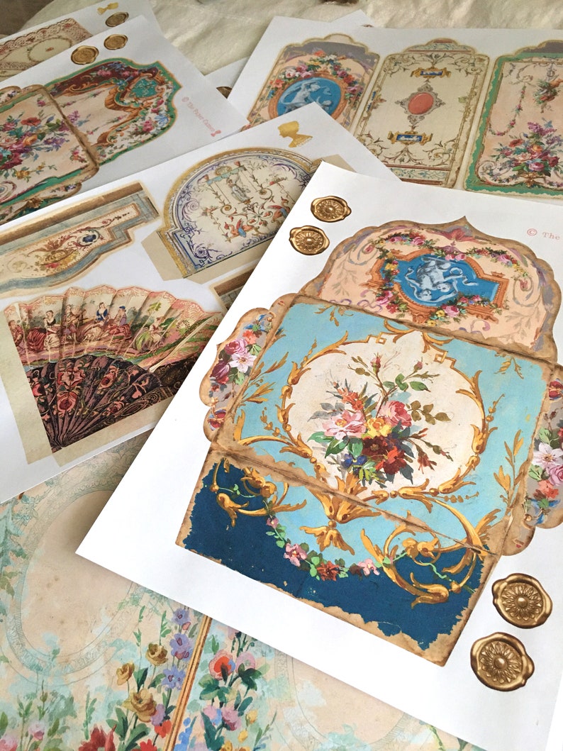 May include: A set of vintage paper ephemera featuring intricate floral designs, gold accents, and a blue and gold patterned box. The paper pieces are printed on a white background and have a distressed, antique look.