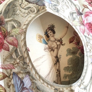 May include: A vintage paper cut-out with a delicate floral design. A woman with a floral crown and butterfly wings is depicted in the center, surrounded by roses and butterflies.