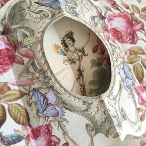 May include: A vintage paper cameo with a delicate floral design featuring roses and butterflies. The cameo features a cutout window revealing a woman with a butterfly on her shoulder.