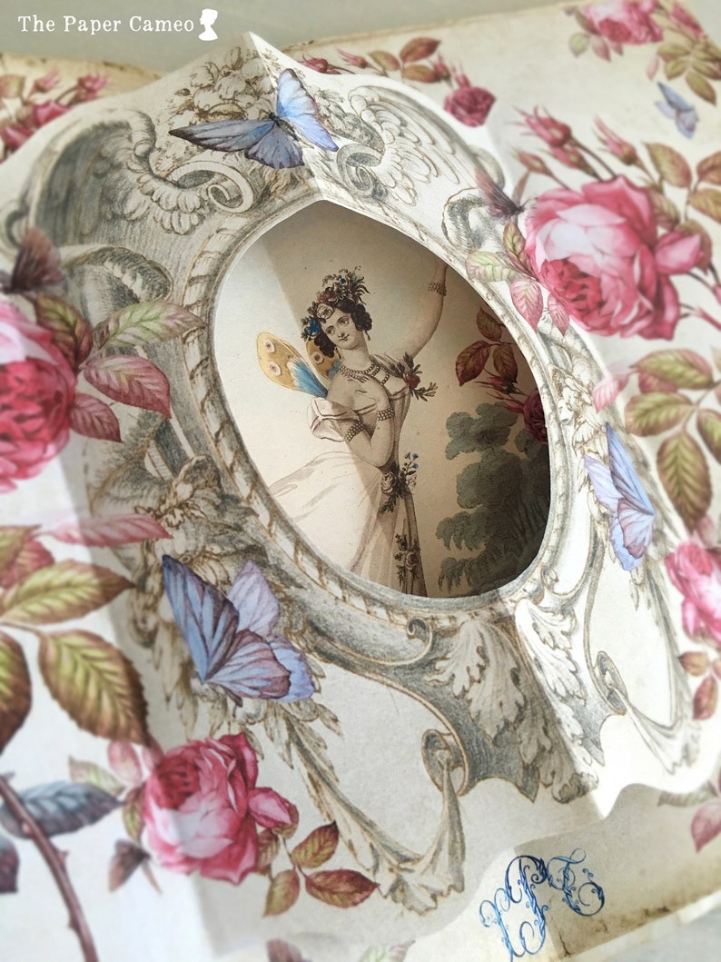 May include: A vintage paper cameo with a delicate floral design featuring roses and butterflies. The cameo features a cutout window revealing a woman with a butterfly on her shoulder.