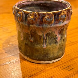 May include: A handcrafted ceramic cup with a distinctive glaze. The cup has a dark brown and green colour scheme, a pinkish-purple rim, and decorative swirls. The glaze has a dripped effect, enhancing its artistic appeal.