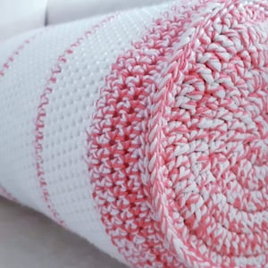 Könnte beinhalten: Ein weißes und rosa gestricktes zylindrisches Kissen mit einem Streifenmuster. Das Kissen ist aus weichem, strukturiertem Garn gefertigt.