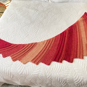 Könnte beinhalten: Ein gehäkeltes Tuch in Rosatönen, Orange und Rot. Das Tuch liegt auf einem weißen Bettbezug.