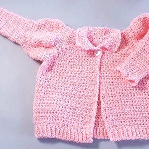 Könnte beinhalten: Eine rosa gehäkelte Strickjacke mit Knopfleiste und Kragen. Die Strickjacke hat lange Ärmel und ein schlichtes Design.