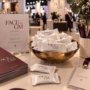 May include: A gold-toned bowl filled with white individually wrapped candies branded with the text FACE by GM AYMIL DESIGN. Nearby are a matching sign, a stack of dark maroon brochures, a white pen holder with pens, and an exhibitor badge.