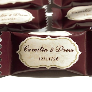 Può includere: Un incarto per caramelle color marrone scuro con bordi seghettati, caratterizzato da un'etichetta color crema decorata al centro. L'etichetta riporta il testo 'Camilia & Drew' in un carattere corsivo marrone, seguito dalla data '12/11/26' in un carattere sans-serif.