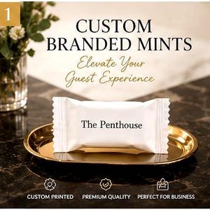 May include: A white rectangular mint wrapper with the text The Penthouse printed in black, resting on a small oval gold-colored metal tray. The image includes text stating CUSTOM BRANDED MINTS, Elevate Your Guest Experience, CUSTOM PRINTED, PREMIUM QUALITY, and PERFECT FOR BUSINESS.